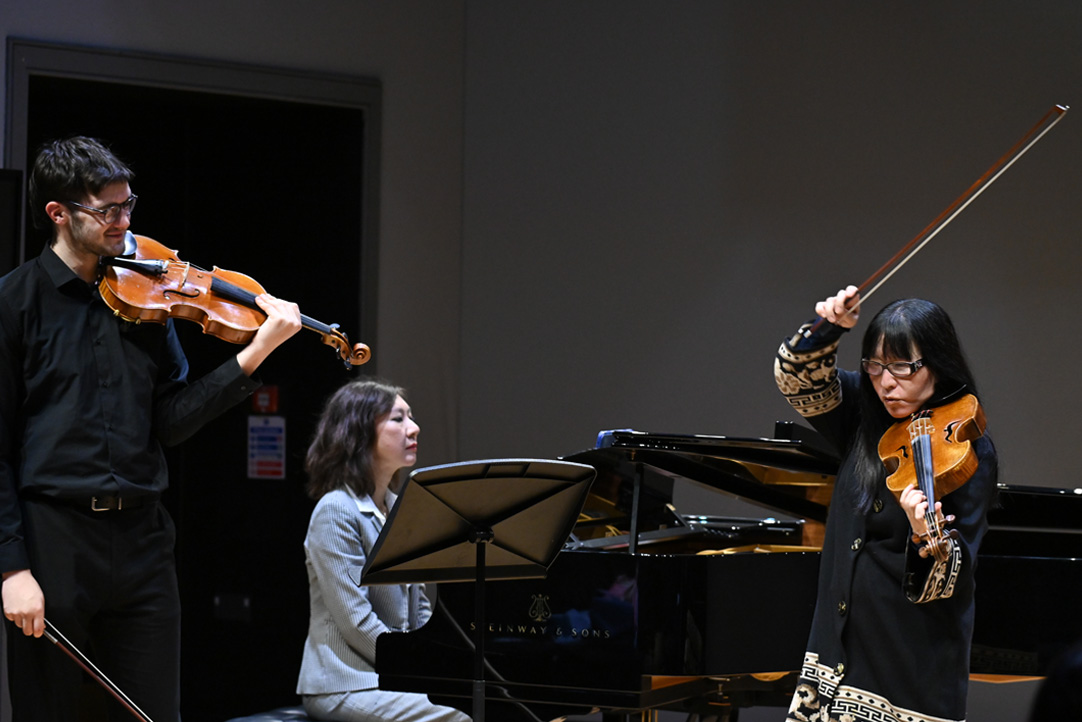 A women playing the viola, with a student holding a viola observing and an accompanist playing.